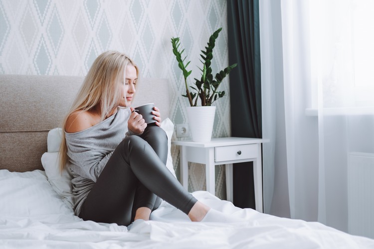 Улыбающаяся красивая белокурая женщина в серой домашней одежде сидит на кровати с чашкой кофе